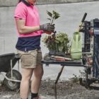 A woman in work clothes holds plants beside gardening tools on a truck tailgate.