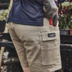 Person in tan work shorts holds red tool with gloved hand; flowers visible in background.