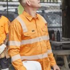 Man in an orange high-visibility shirt holding a hard hat and thermos, looking upward.