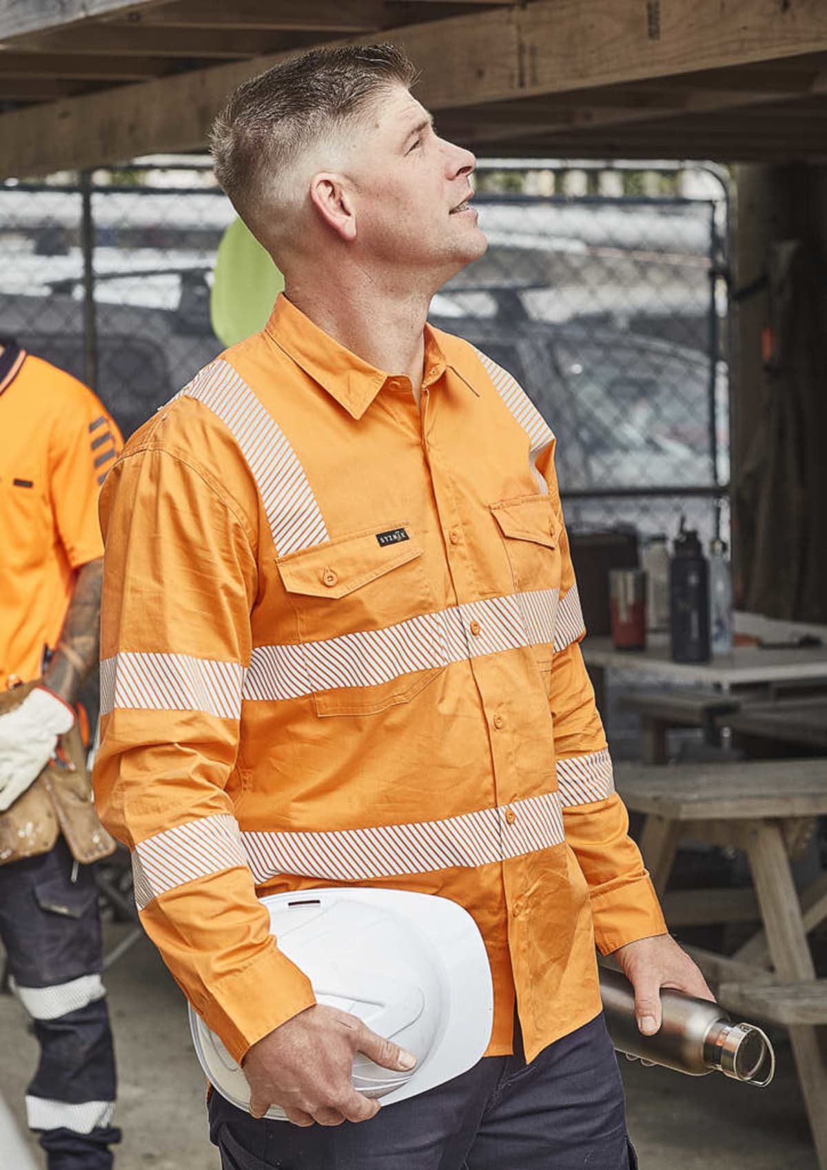 Man in an orange high-visibility shirt holding a hard hat and thermos, looking upward.