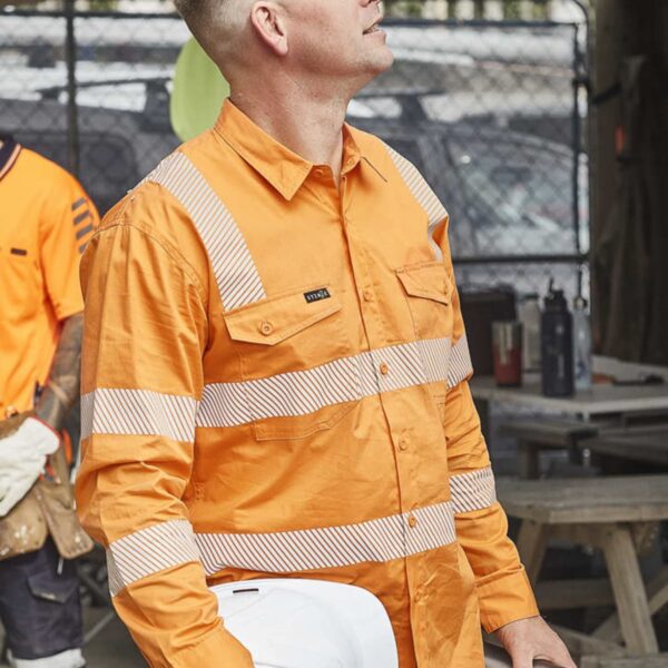 Man in an orange safety shirt holds a hard hat and thermos, looking up at a construction site.