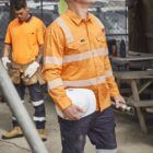Two construction workers in orange safety shirts stand at a job site, one holding a hard hat.