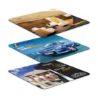 Three rectangular mouse pads in perspective view. Top pad depicts a modern office lounge; middle pad features a blue car; bottom pad shows a hand holding a small house model. Each pad has colorful branding and text.
