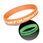 An orange wristband with STAR FOUNDATION printed in white. Below it, a green version glows in the dark, showing the same text.