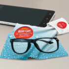 Smartphone, black-framed glasses, and two screen cleaning cloths (one blue with water droplets, one white with a red logo) on a gray surface.