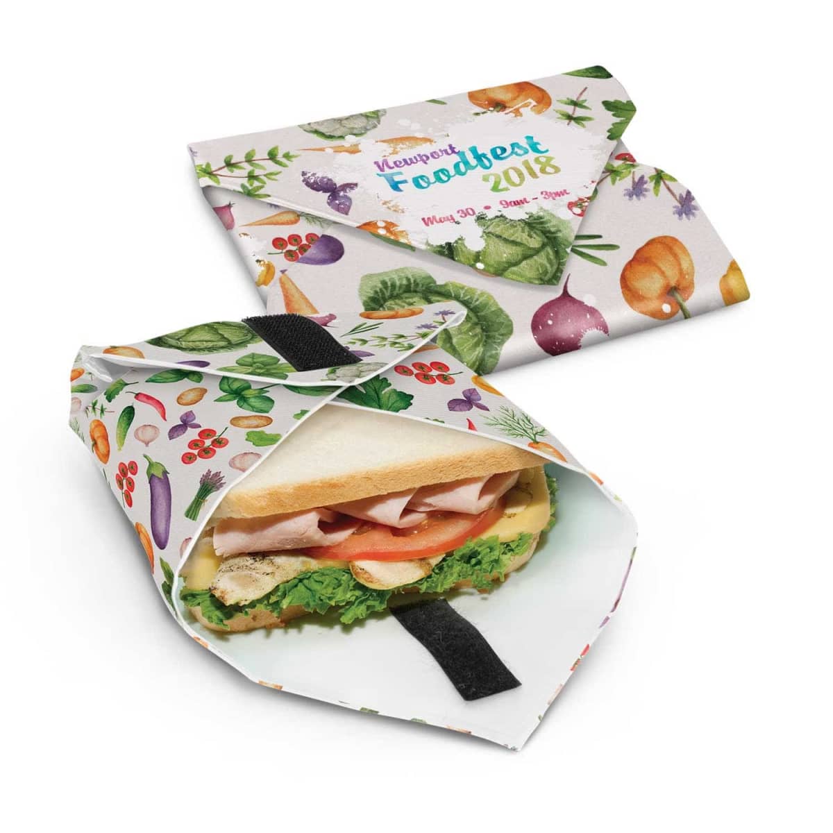A sandwich with lettuce, tomato, and turkey is partially wrapped in a reusable, colorful sandwich wrap featuring various fruits and vegetables. Another closed wrap lies beside it, displaying Newport Foodfest 2018 with matching fruit and vegetable patterns.