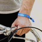 Close-up of a person holding bicycle handlebars with their right hand. The person is wearing a blue Triathlon wristband. They are dressed in a black and white striped shirt.