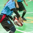 A child in a blue jersey and black shorts is holding a rugby ball on a green indoor field. Other players in similar attire are visible in the background.