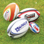 Four colorful rugby balls with different designs and logos are placed on a grassy field. The brands visible are Creatorex, WoodZilla, Minnan Strategy Solutions, and one with a red, white, and blue pattern.