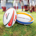 Two rugby balls are on the grass, one with red and gray designs labeled AB Sports, and the other with blue and yellow stripes labeled Southern Rugby League. Blurred players in red uniforms are in the background.