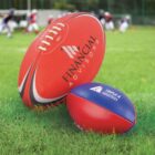 Two rugby balls on grass: a larger red ball with Financial Advisors text and a smaller blue and red ball with Triple A Industries text. Blurred background shows people playing rugby.