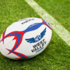 A rugby ball with red and blue designs and the words RugbyEye and West Valley Tours lies on a grassy field near a white line.