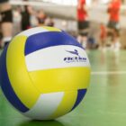 A volleyball with yellow, white, and blue panels sits on a green indoor court. The words Active sport & fitness are visible on the ball. Blurred players in red and black uniforms are in the background.