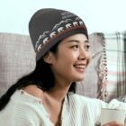 A woman is smiling while sitting indoors, wearing a gray beanie with a mountain and bear pattern. She is holding a white mug and dressed in a white knit sweater. Behind her is a plaid blanket draped over a chair.
