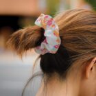 Close-up of a persons hair tied in a ponytail with a colorful scrunchie. The scrunchie has patterns of different icons, including fruits and geometric shapes, on a light background. The person is outside with a blurred building in the background.