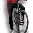 A cyclist wearing black and red athletic clothing is on a mountain bike, positioned with one foot on the pedal and the other on the ground. The background is a simple white to gray gradient.