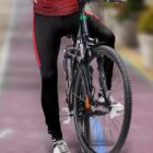 Person wearing a red jersey and black leggings, riding a mountain bike on a paved path. The bike has a black frame with green and orange accents. The riders face is not visible.