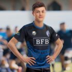 A rugby player in a blue and black jersey with BFB and RUGBY logos stands confidently with hands on hips. The background is blurred, suggesting a sports setting with other players or a field.
