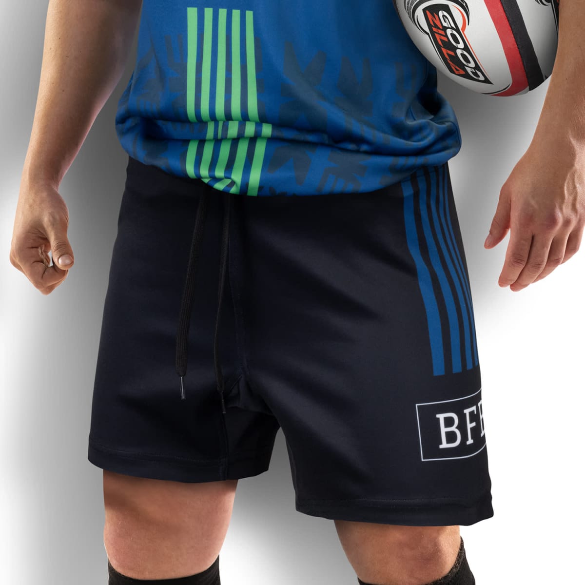 Person wearing a blue and green sports jersey and black shorts holds a rugby ball in one arm.