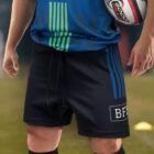 A person in a blue and green sports jersey and black shorts, holding a rugby ball tucked under one arm. The shorts have blue stripes and a BFF logo. The person is standing outdoors on a grassy field.