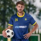 A soccer player in a blue and yellow team jersey holds a soccer ball. The jersey features the number 26 and a logo with the word CONNECT. Blurred greenery is in the background.