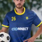 A soccer player in a blue and yellow team jersey holds a soccer ball. The jersey features the number 26 and a logo with the word CONNECT. Blurred greenery is in the background.