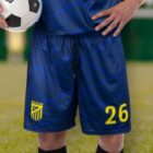 A soccer player holding a ball stands on a grass field, wearing a blue uniform with yellow number 26 and a yellow and blue crest on the shorts. Only the lower half of the players body is visible.