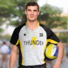 A volleyball player in a Thunder team jersey holds a volleyball. The jersey is gray with black and yellow accents and features the number 18. The background is blurred with greenery and people.