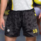 A person wearing black and gray camo-patterned sports shorts with the number 18 and a logo. Theyre holding a yellow and blue ball. The background is blurred, focusing on the shorts and ball.