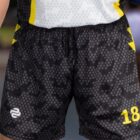 A person wearing black and gray camo-patterned sports shorts with the number 18 and a logo. Theyre holding a yellow and blue ball. The background is blurred, focusing on the shorts and ball.