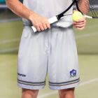 A person in a light grey tennis outfit holds a racket and a yellow tennis ball on a court. Their shorts have a striped trim with a small emblem on the right leg. The background features a blurred green surface and netting.