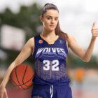 A basketball player in a Wolves jersey, numbered 32, holds a basketball under one arm while giving a thumbs up with the other hand. The player has their hair tied up and stands on an outdoor court with a blurred background.