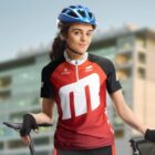 A woman in a red and black cycling jersey and blue helmet stands with a bicycle. She has a confident expression and is wearing cycling gloves. A modern building with glass windows is blurred in the background.