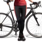 A person in athletic gear leans against a road bike. They wear a red jersey, black gloves, black and red cycling pants, and black shoes. The bike is a sleek black model with drop handlebars, set against a plain white background.