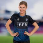 A person in a dark blue sports jersey with various logos, including BFB, smiles with hands on hips against a blurred outdoor background. The jersey features geometric patterns with red details.