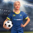 A woman with blonde hair in a ponytail, wearing a blue and yellow soccer jersey with the number 26, smiles while holding a soccer ball under her arm. She stands in a stadium filled with blurred lights in the background.