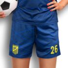 A person in a blue sports uniform with geometric patterns holds a soccer ball. The number 26 is printed in yellow on the shorts, along with an emblem. The persons fingernails are painted red.