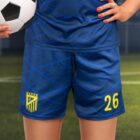 A person holding a soccer ball is wearing a blue soccer uniform with the number 26 and a yellow crest on the shorts. The uniform has a subtle geometric pattern. The background features a blurred soccer field.