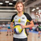 A female volleyball player in a THUNDER jersey, holding a volleyball, stands on an indoor court. Other players are blurred in the background. The setting is a large gymnasium with bright lights.