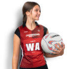 A netball player in a red and black uniform holds a netball. Her jersey displays CHAMPIONS and WA. She is smiling and looking to the side.
