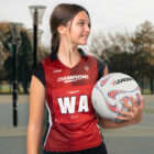 A young woman stands on an outdoor netball court, holding a netball. She is wearing a red and black netball uniform with CHAMPIONS and WA on the bib. The background shows netball hoops and a blurred view of trees.