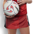 A person wearing a red and black striped sports uniform holds a white and multicolored netball labeled LEADER. Their face is not visible, and the background is plain white.