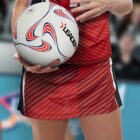 A person in a red and black striped sports uniform holds a white netball with blue and red patterns. The word LEADER is visible on the ball. The background is blurred, suggesting an indoor court setting.