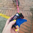 A hand with yellow-painted nails holds a colorful pouch containing blue dog waste bags. A small dog is visible in the background walking on a brick path with a pink leash.