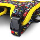 Close-up of a colorful pet harness with a yellow trim. It features a black plastic buckle and a pattern of various cartoon symbols and emojis on a dark background.