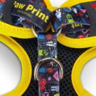 Close-up of a colorful dog harness with vibrant patterns and text, including Paw Print and GROOMING. The harness features yellow and black fabric with metal rings and a padded interior.