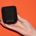 Hand holding a black portable speaker with buttons, against an orange background.