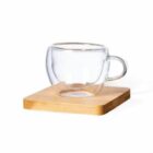 Clear double-walled glass cup on a wooden coaster, isolated on a white background.