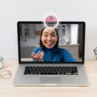 Woman in blue sweater video calling on a laptop with a ring light attached.