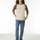 Person in a beige and brown raglan shirt, blue jeans, and white sneakers stands facing forward.